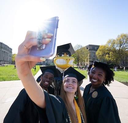 UI students in graduation apparel take a selfie photo on the UI Pentacrest