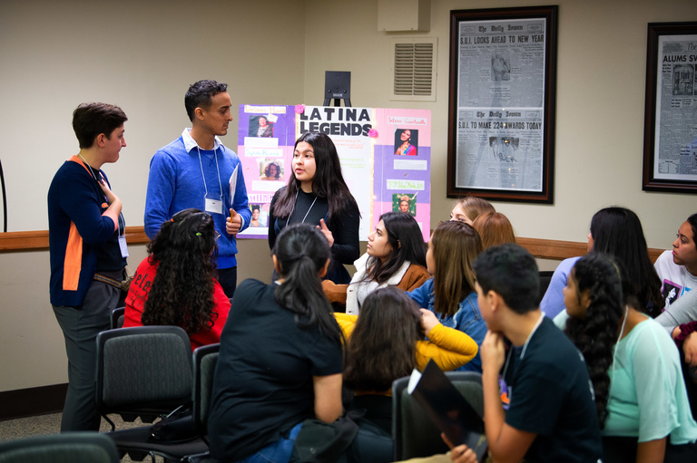 Group of students look at 3 speakers with a "Latina Legends" informational poster behind them